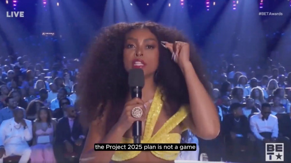 Image of Taraji P. Henson hosting the 2024 BET Awards. She has long, brown, curly hair and is dressed in a lovely and fancy looking yellow top. She is speaking directly to the camera with the audience visible behind her and the caption underneath her says "the project 2025 plan is not a game."