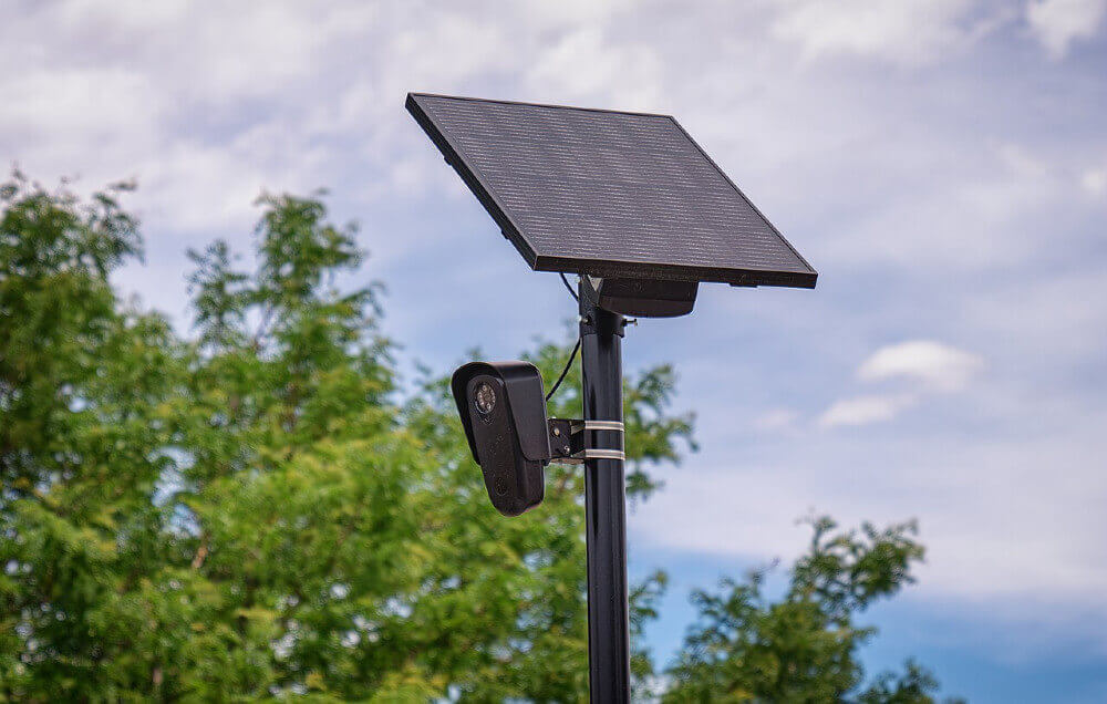 Photograph of a Flock Safety camera. There are trees and sky in the background, and there is a black metal pole with a small camera mounted on it, and at the top is a small solar panel mounted on it.