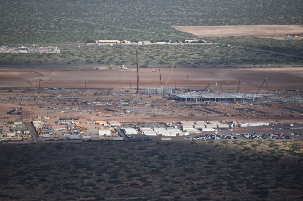 Aerial view of Project Jupiter, a large data center construction project in progress with hundreds of workers, cars, and several cranes in the distant. It is a desert landscape in the background, in Southern New Mexico. 