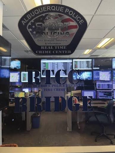 Image of a glass door with a large Albuquerque Police Real Time Center sticker on it and text that says RTCC Bridge. Through the glass there are large screens with data on them, surrounded by command center style chairs. 